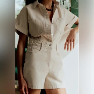 Beige heans jumpsuit Zara  - Snygga beige shorts med hög midja och klassisk femficksdesign. De är tillverkade i ett jeansliknande material och har bälteshällor samt knapp- och dragkedjestängning. Perfekta för en clean och trendig look i sommar.