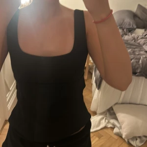 Svart och vit korsettopp med rosett - Två stilrena korsettoppar i svart och vitt med breda axelband och fyrkantig urringning. Baksidan har en snygg öppen detalj med stor rosett som knyts. Topparna är figurnära och har en clean, modern look som passar perfekt till jeans eller kjol.