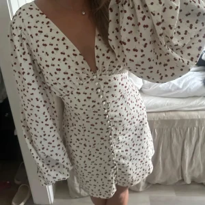 Vit kortklänning med körsbärsmönster - Supersöt vit kortklänning med röda körsbär, djup v-ringning och stora puffärmar. Klänningen har knappar framtill. Perfekt för sommarens alla tillfällen!