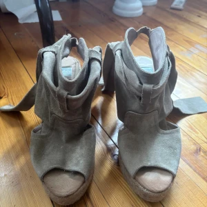 Beiga mockasandaler med kilklack - Snygga sandaler från Kanna i beige mocka med öppen tå och knytband runt ankeln. Skorna har en hög kilklack i flätad jute som ger en cool espadrillekänsla. Perfekta för dig som vill ha både höjd och komfort i sommarens outfits. En söm som har gått av på ena men de går att fixa.            Priset+frakt