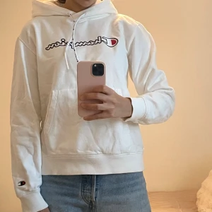 Vit hoodie från Champion med logga - Vit hoodie från Champion med broderad logga över bröstet och liten logga på ärmen. Klassisk känguruficka framtill, dragsko i huvan och mjukt bomullsmaterial. Perfekt för chill dagar eller när du vill ha en clean streetstyle.