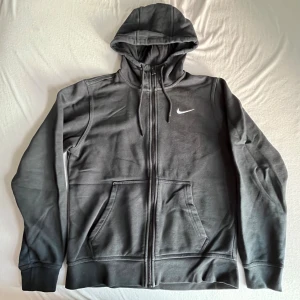Svart Nike hoodie med dragkedja - Svart hoodie från Nike med vit logga på bröstet. Tröjan har huva med snörning, hel dragkedja och två stora fickor framtill. Tillverkad i mjuk bomullsblandning, perfekt för chill eller träning. Klassisk och enkel design som funkar året runt.