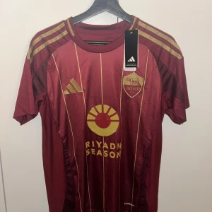 AS Roma Dybala matchtröja Adidas S - AS Roma fotbollströja från Adidas med Dybala 21 på ryggen. Tröjan är vinröd med guldiga detaljer, smala vertikala ränder och klubbmärke på bröstet. Tillverkad i lätt funktionsmaterial (AEROREADY) och har korta ärmar. Perfekt för dig som älskar Serie A och snygg design.