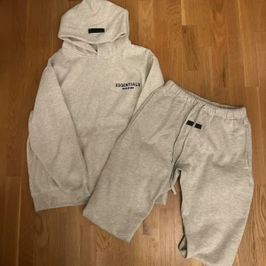 Grå Essentials Fear of God hoodie - Säljer en ljusgrå hoodie från Essentials Fear of God med svart logga på bröstet och ryggen. Tröjan har huva, ribbade muddar och är tillverkad i mjuk bomullsmix. Perfekt för en chill och stilren look.