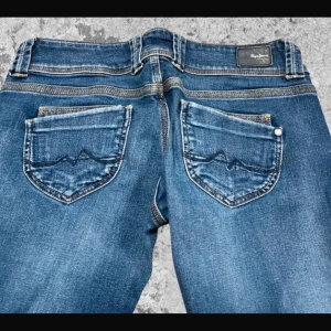 Blå bootcut jeans från Pepe Jeans - Snygga blå jeans från Pepe Jeans med bootcut passform och klassisk femficksdesign. Jeansen har dubbla knappar i midjan, raka bakfickor med vågig brodyr och lätt slitna detaljer. Perfekta för dig som gillar en retroinspirerad look.