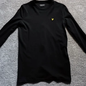 Svart långärmad tröja Lyle & Scott - Svart långärmad tröja från Lyle & Scott med klassisk gul logga på bröstet. Enkel och stilren design, perfekt till jeans eller joggers. Mjuk och skön bomullskvalitet som passar året runt.