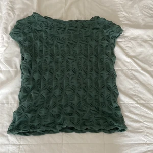 Grön strukturerad topp från Lindex - Säljer en grön topp från Lindex i storlek S. Toppen har ett unikt strukturerat mönster över hela tyget och är kortärmad med vågiga kanter. Perfekt för dig som vill ha något annorlunda och snyggt i garderoben.