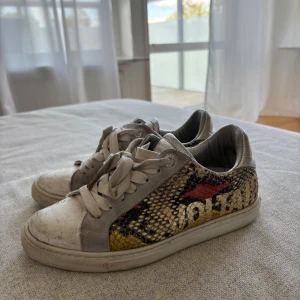 Zadig & Voltaire sneakers  - Coola sneakers från Zadig & Voltaire med ormmönstrade sidor i beige, svart, gul och röd. Skorna har grå mockadetaljer, vita snören och platt sula. Märkeslogga på hälen och sidan. Perfekta för dig som vill sticka ut med en edgy streetstyle.