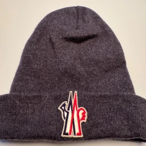 Mörkgrå Moncler mössa i ull - Snygg mörkgrå mössa från Moncler med broderad logga framtill i rött, vitt och blått. Tillverkad i mjuk ull som håller dig varm under kalla dagar. Klassisk uppvikt kant och stilren design som passar perfekt till vintern. Tophat / beanie 