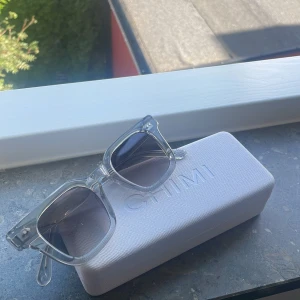 Transparenta solglasögon från CHIMI - Snygga solglasögon från CHIMI med transparent, fyrkantig båge och mörktonade glas. Riktigt clean och modern look som passar perfekt till sommaren. Kommer med originaletui med CHIMI-logga. Perfekt accessoar för att lyfta din stil. 04or 