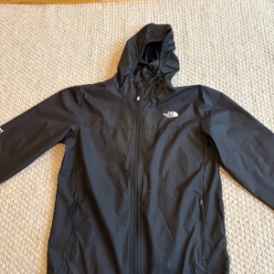  North Face jacka  - Svart vindjacka från The North Face i storlek XL. Den är mindre i storleken så passar L och även M. Jackan har huva, hel dragkedja framtill och två sidofickor med dragkedja. Snygg logga på bröstet och ryggen. Perfekt för blåsiga dagar och outdoor-aktiviteter.