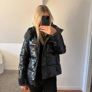 Calvin Klein High Shine Puffer Jacket - Snygg svart pufferjacka från Calvin Klein med glansig finish och huva. Jackan har CK-logga på bröstet och Calvin Klein-text på ärmen. Dragkedja framtill samt knappar att stänga med. Jackan är i riktigt bra skick då jag nästan aldrig använt den. Är i storlek M men har själv S i vanliga fall så den är lite mindre i storleken. Är ca 170 cm lång💞