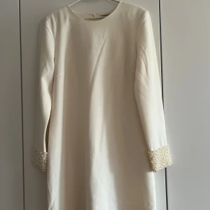 Vit långärmad klänning med pärlor - Superfin vit klänning med lång ärm och rund halsringning. Ärmsluten är dekorerade med massor av pärlor som ger en lyxig och unik touch. Klänningen har en rak och enkel passform, perfekt för dig som gillar stilrena plagg med det lilla extra.