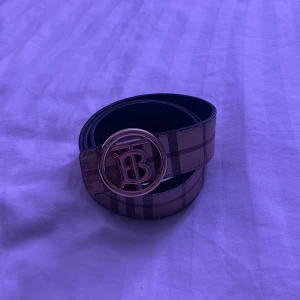 Burberry vändbart bälte med logga - Snyggt vändbart bälte från Burberry med klassiskt rutigt mönster på ena sidan och svart skinn på andra. Stort, rundat spänne i guld med TB-logga. Perfekt accessoar för att lyfta din outfit med lyxig känsla och ikonisk design. Lite använd vid hålen men märks inte.