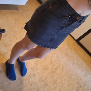 Svart jeanskjol från 157 - Snygg svart jeanskjol från 157 med rå kant nertill och klassiska fickor både fram och bak. Kjolen har en enkel knappstängning med flera knappar som jylf. Perfekt för dig som gillar en stilren och edgy look. Det står XS mem jag skulle mer säga att det är en S eller M