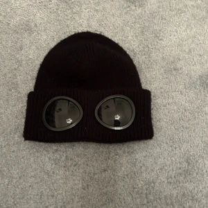 Svart C.P. Company goggle-mössa ej äkta - Svart stickad mössa från C.P. Company med deras ikoniska goggle-linser framtill på uppviket. Mössan är ribbstickad och har en klassisk form som sitter snyggt på huvudet. Perfekt accessoar för att sticka ut i mängden.