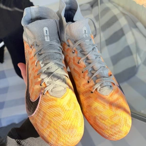 Nike super fly 9 Elite - Snygga orangea fotbollsskor från Nike med grå snörning och svart Nike-logga. Skorna har en hög ankelkrage för extra stöd och är perfekta för fotbollsplanen.