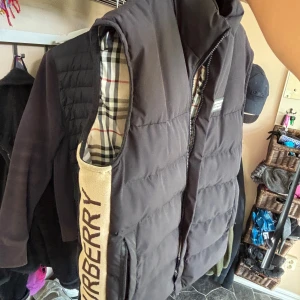 Svart dunväst från Burberry  - Svart dunväst från Burberry med klassiskt rutigt foder i beige, svart och rött. Västen två fickor med dragkedja och Burberry-logga på bröstet. Stängs med dragkedja framtill och har en loose passform.