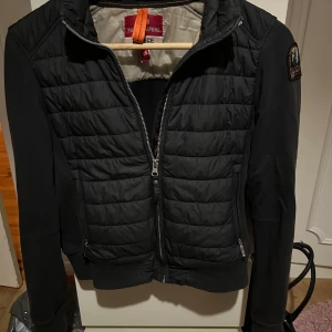 Svart quiltad jacka Parajumpers XS - Svart quiltad jacka från Parajumpers med ribbade muddar, dragkedja framtill och två sidofickor med dragkedja. Jackan har lång ärm, patch på ena ärmen och en stilren look. Perfekt för dig som gillar streetwear och vill ha något snyggt och funktionellt.