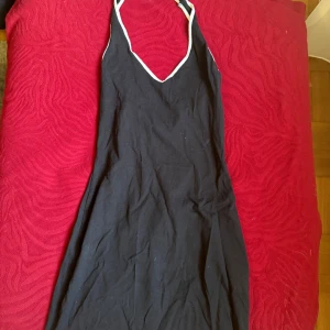 Svart kortklänning från H&M Divided - Svart kortklänning från H&M Divided med vita kanter och smala axelband. Klänningen har en v-ringad hals och är ärmlös, perfekt för varma dagar. Tillverkad i mjuk bomull som känns skön mot huden.