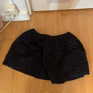 Svart broderad kjol från H&M - Svart kortkjol från H&M med broderade detaljer och genombrutet mönster. Kjolen har resår i midjan och är fodrad på insidan för extra komfort. Perfekt för dig som gillar en söt och trendig stil.