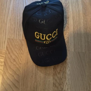 svart gg keps - Snygg svart keps från gg med broderad logga i gult, grönt och rött framtill. Kepsen har klassisk böjd skärm och diskret mönster med gg-tryck över hela tyget. Perfekt accessoar för att lyfta din outfit.