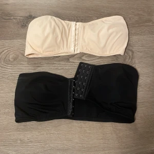 2-pack bandeau tubtoppar svart & beige - Säljer två bandeau tubtoppar i svart och beige. Båda är axelbandslösa med vaddering och har hakknäppning på framsidan. Tillverkade i mjukt syntetmaterial som sitter skönt mot huden. Aldrig använda