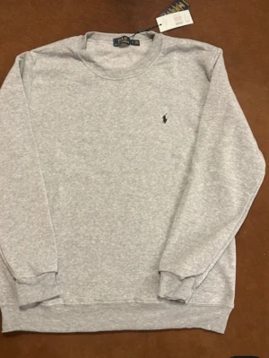 Grå sweatshirt från Polo Ralph Lauren - Klassisk grå sweatshirt från Polo Ralph Lauren med rund halsringning och diskret broderad logga på bröstet. Tillverkad i mjuk bomullsmix och har ribbade muddar vid ärmslut och nederkant. Perfekt för en avslappnad och stilren look.