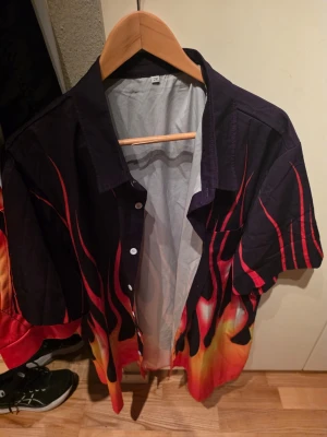 Svart skjorta med eldflammor 2XL - Säljer en kortärmad svart skjorta i storlek 2XL med fett eldflammigt tryck i rött, orange och gult. Skjortan har klassisk krage, vita knappar och en bröstficka. Perfekt för dig som vill sticka ut och köra på en riktigt cool look.