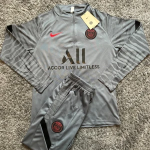 Grå PSG x Nike träningsset - Säljer ett grått träningsset från Nike x Paris Saint-Germain med långärmad tröja och byxor. Setet har svarta och röda detaljer, PSG-logga på bröst och ben, samt halv dragkedja på tröjan. Tillverkat i mjuk polyester med diskret mönster på ärmar och ben. Pris går självklart att diskutera!