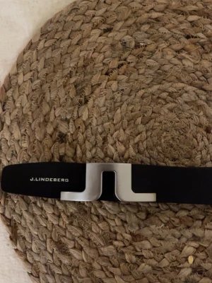 Svart bälte från J.Lindeberg - Säljer ett svart bälte från J.Lindeberg med en stilren, matt silverfärgad metallspänne i form av ett J. Bältet har en modern och minimalistisk look som passar till många olika outfits.