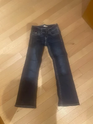 Mörkblå bootcut jeans med kontrastsömmar - Säljer ett par mörkblå jeans med tydliga vita kontrastsömmar och snygga fickdetaljer bak med lock och knapp. Klassisk femficksmodell och bootcut-ben som ger en schysst siluett. Perfekta till sneakers eller boots.