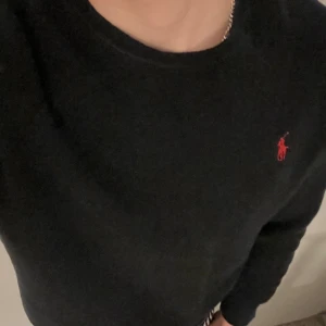 Svart tröja från Polo Ralph Lauren - Klassisk svart tröja från Polo Ralph Lauren med rund hals och den ikoniska röda loggan broderad på bröstet. Tröjan är långärmad och gjord i mjuk bomull, perfekt för en clean och stilren look.