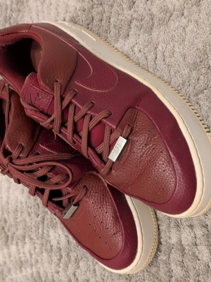 Vinröda Nike Air Force 1 sneakers - Säljer ett par vinröda Nike Air Force 1 sneakers i skinn med klassisk vit sula. Skorna har rund tå, snörning med metall-detaljer och den ikoniska swoosh-loggan på sidan. Perfekta för dig som vill ha en clean och stilren look med en twist.