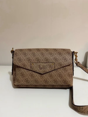 Guess väska  - Brun axelväska från Guess med logomönster över hela väskan. Väskan har guldfärgade metalldetaljer, justerbar axelrem och flera fack inuti. Stängs med magnetknapp och har dragkedja på baksidan. Dragkedjan på baksidan har inte en dragare men det funkar ändå!