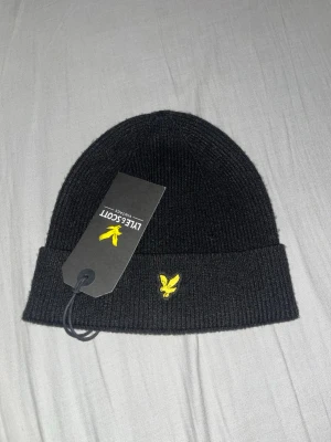 Svart mössa från Lyle & Scott - Säljer en svart ribbstickad mössa från Lyle & Scott med gul broderad logga framtill. Klassisk uppvikt kant och mjukt material som sitter skönt på huvudet. Perfekt accessoar för kyliga dagar.