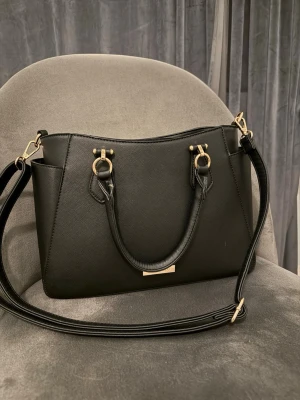 Svart handväska från Anna Field - Stilren svart handväska från Anna Field med guldfärgade metalldetaljer och strukturmönstrad yta. Väskan har både handtag och justerbar axelrem, perfekt för dig som vill ha en snygg och praktisk väska till exempelvis skolan. Praktisk innerficka. Mycket fint skick på väskan