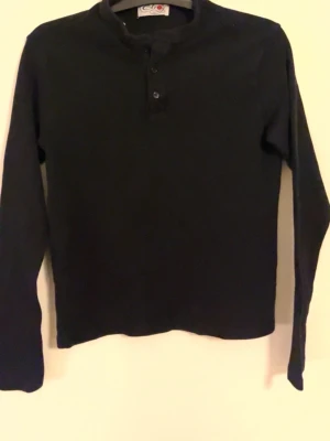 Svart långärmad pikétröja Elroy - Svart långärmad pikétröja från Elroy med knappar framtill och en liten ståkrage. Tröjan har en enkel och clean look med diskret broderad logga på ryggen vid axeln. Perfekt till chill vardagsstil. 