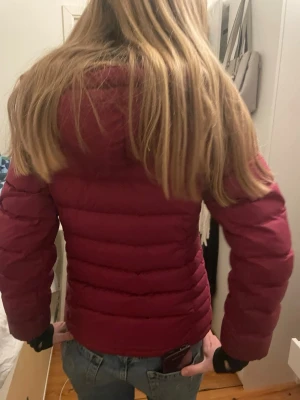 Vinröd dunjacka från bergans bra för skidåkning  - Säljer en vinröd dunjacka från bergans med svarta dragkedjor och två sidofickor. Jackan är quiltad och har en smal passform som sitter snyggt. Perfekt för kalla dagar och enkel att matcha med jeans eller hoodies.