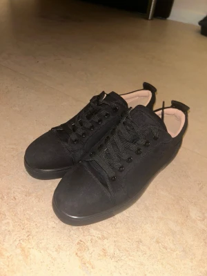 Svarta Christian Louboutin utan spikar - Snygga svarta sneakers från Christian Louboutin i mocka med klassiska röda sulor och broderad logga bak. Skorna har rund tå, svarta snören och beige innersula med guldfärgad logotyp. Perfekta för dig som vill ha en lyxig touch till din outfit. 