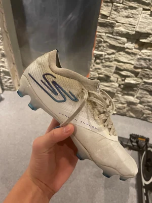 Vita Skechers razor - Säljer ett par vita Skechers razor fotbollsskor med blå detaljer och snörning. Skorna har en lätt ovandel i syntetmaterial och mönstrad sula med blå dobbar för bra grepp på planen. Texten 'Carbon Infused' syns på sidan och designen är sportig och modern.