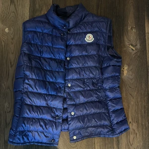 Blå/lila dunväst från Moncler - Snygg blå dunväst från Moncler med knappar framtill och ett stilrent märke på bröstet. Perfekt för kyligare dagar när du vill hålla stilen. Västen har en hög krage för extra värme. Storlek 1/S