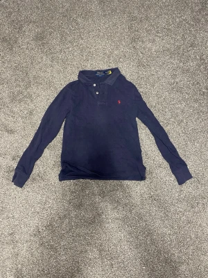 Mörkblå tröja från Polo Ralph Lauren - Mörkblå långärmad tröja från Polo Ralph Lauren med slim fit. Skjortan har klassisk krage, knappar framtill och den ikoniska röda broderade loggan på bröstet. Tillverkad i mjuk bomull som känns skön mot huden.