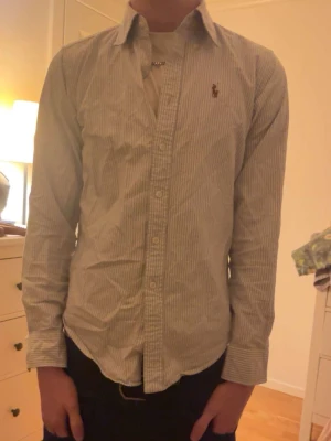 Randig skjorta från Ralph Lauren - Klassisk skjorta från Ralph Lauren med tunna vertikala ränder i vitt och ljusblått. Skjortan har lång ärm, normal passform och knappar framtill. Ikoniska Polo-loggan broderad på bröstet. Perfekt till jeans eller chinos.