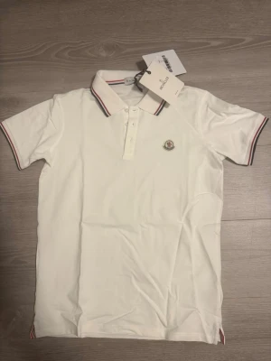 Vit Moncler pikétröja med ränder - Klassisk vit pikétröja från Moncler med korta ärmar och krage. Tröjan har diskreta röda och marinblå ränder på krage och ärmslut samt Moncler-logga broderad på bröstet. Tillverkad i mjuk bomull för en bekväm känsla scan finns.