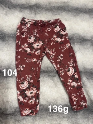 Blommiga mjukisbyxor i vinrött - Säljer ett par vinröda mjukisbyxor med blommigt mönster. Byxorna har resår i midjan och dragsko för skön passform. Perfekta för chill dagar eller när du vill ha något bekvämt och snyggt på dig.🌸 Fint använt skick. Storlek 104