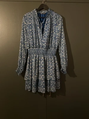 Blåvit kortklänning med mönster - Supersnygg blåvit kortklänning från Zara med grafiskt mönster och smock i midjan. Klänningen har lång ärm, v-ringning och volangdetaljer i kjolen. Säljs inte längre och är använd 1 gång!