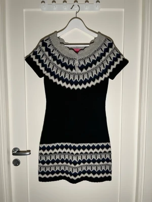 Stickad kortklänning från Indiska - Mysig stickad kortklänning från Indiska i svart, grått och marinblått. Klänningen har korta ärmar, rund halsringning med tre knappar och ett snyggt grafiskt mönster upptill och nedtill. Perfekt för lager på lager under höst och vinter.