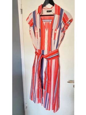 Randig klänning - En luftig klänning från Lindex i rött blått och vitt. Med klänningen kommer ett tjockt band man kan knyta i midjan, bandet är inte fastsytt. Plagget har två bröstfickor med knappar samt knappar i mitten. På sidan av klänningen finns det en dragkedja (se på sista bilden).  Material: 100% bomull 