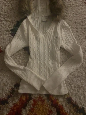 Vit stickad hoodie från Hollister XS - Säljer en vit kabelstickad hoodie från Hollister i storlek XS. Tröjan har v-ringning, långa ärmar, ficka framtill och huva med fuskpälsdetalj. Klassisk Hollister-logga broderad på bröstet. Perfekt för kyliga dagar! Så fin och ger verkligen 2000s vibes! Jag är öppen för prisförslag. Men inte överdrivet lite.                                                               Högsta budet just nu är 699kr! Buda högre än det så kanske just du får tröjan! 🤍